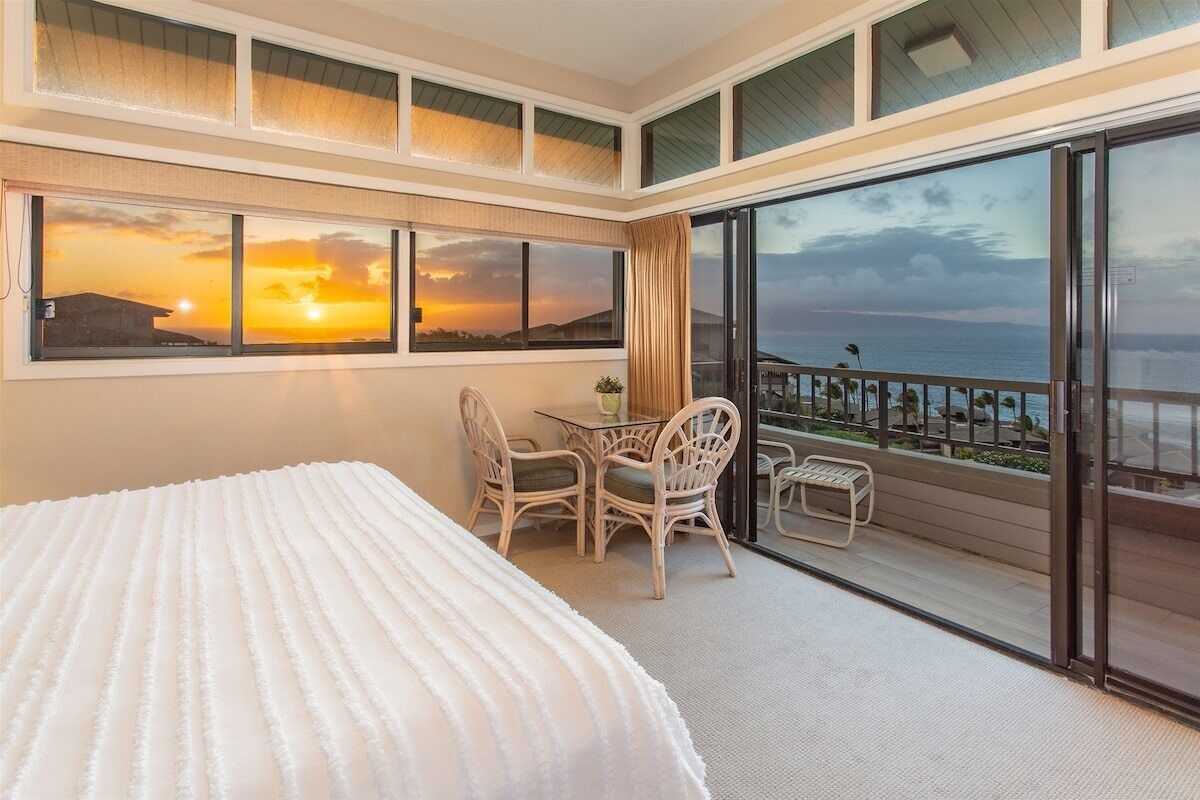 Une chambre d’hôtel avec un grand lit, un balcon doté d’une table et de chaises, et une vue imprenable sur le coucher de soleil.