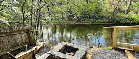 A wooden deck with a stone fire pit and benches overlooking a calm lake surrounded by trees.