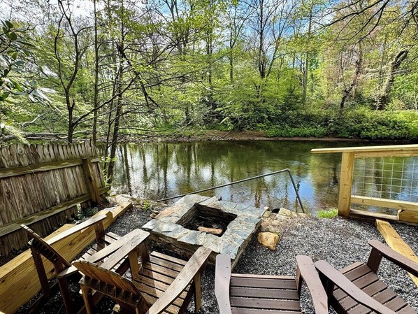 A wooden deck with a stone fire pit and benches overlooking a calm lake surrounded by trees.