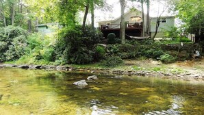 A house with a deck overlooking a clear stream, surrounded by dense greenery. - Rivers Edge Cabin, Swimming, Tubing, Fishing, Fire Pit, Grill, Wi-Fi, Relaxing! (Burnsville)