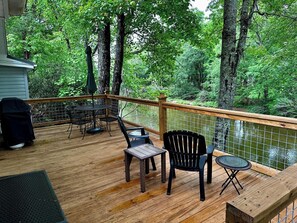A wooden deck with outdoor furniture, a view of a forest, and a water feature. - Cabin on the River+Fire Pit+Grill, WiFi, Private! (Burnsville)