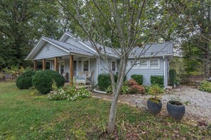 A two-story house with a porch, surrounded by a well-maintained garden with various shrubs and a tree. - Cabin on the River+Fire Pit+Grill, WiFi, Private! (Burnsville)