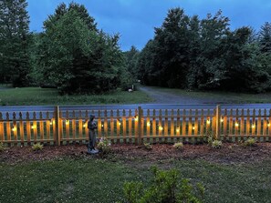Una valla de madera con luces de cuerda, una estatua y una zona cubierta de césped con árboles al fondo.