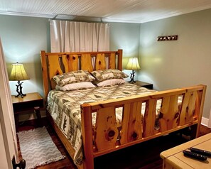 A wooden bed with a quilt, two bedside lamps, and a small table with a remote control. - Cabin on the River+Fire Pit+Grill, WiFi, Private! (Burnsville)