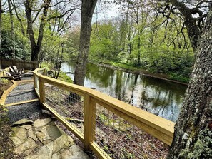 A wooden deck with a railing overlooking a calm body of water, surrounded by trees. - Cabin on the River+Fire Pit+Grill, WiFi, Private! (Burnsville)