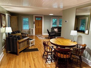 A room with a wooden floor, a dining table with chairs, a sofa, and a door. - Cabin on the River+Fire Pit+Grill, WiFi, Private! (Burnsville)
