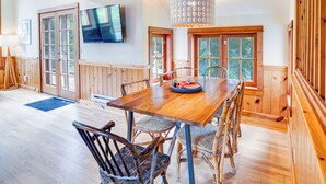 A dining area with a wooden table and chairs, a television mounted on the wall, and large windows with a view of the outdoors. - River to Breakers - River View (Yachats)