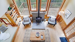 A cozy living room with a fireplace, wooden furniture, and large windows overlooking a deck. - River to Breakers - River View (Yachats)