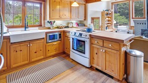 A wooden cabin kitchen with a large window, wooden cabinets, a stove, and a sink. - River to Breakers - River View (Yachats)
