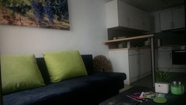 Una sala de estar con un sofá azul oscuro, cojines verdes, una mesa de centro con objetos desordenados y una zona de cocina con armarios y una encimera.