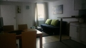 Una habitación pequeña y tenuemente iluminada, con kitchenette, un sofá con cojines verdes y una mesa de comedor con sillas.