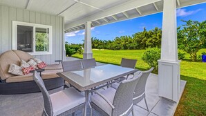 Eine überdachte Terrasse mit Glastisch und Korbstühlen, mit Blick auf einen gepflegten Rasen und einen Golfplatz.