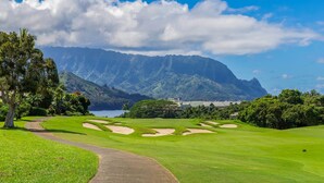 Ein Golfplatz mit Bunkern, einem Fußweg und Blick auf Berge und ein Gewässer.