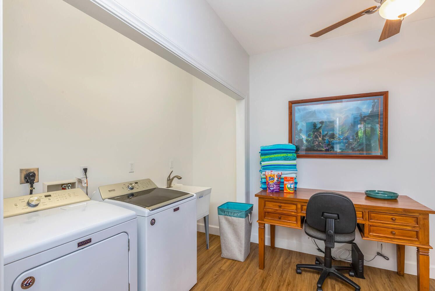 A laundry room with a washing machine, dryer, and a desk with a chair. There is a painting on the wall and a small table with laundry supplies.