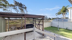 Une terrasse couverte avec un barbecue, une clôture blanche et un arbre en arrière-plan.