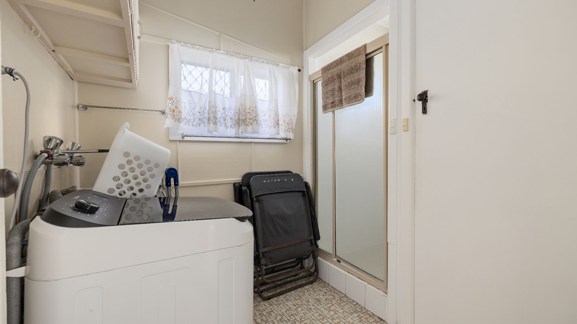 Ein kleines Zimmer mit Waschmaschine, einem Stuhl, einem Koffer und einem Fenster mit Vorhang.