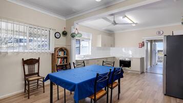 A dining area with a blue tablecloth, wooden chairs, a bookshelf, and a kitchen with white cabinets and a stove.