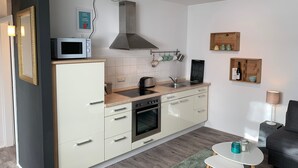 A modern kitchen with white cabinets, a built-in oven, a microwave, and a stainless steel range hood. There is a small island with a sink, and a wooden shelf above the sink with kitchenware. - Cozy 2 room apartment in Mainzer Bestlage (Oberstadt)