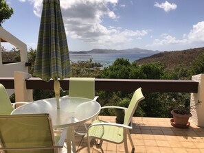A terrace with a glass table and chairs, a striped umbrella, and a view of the sea and hills. - Guavaberry Hill Villa - Amazing sunset views by National Park, No A/C (St. John)