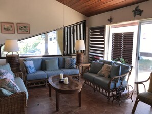 A cozy living area with a blue sofa, wicker chairs, a wooden coffee table, and a view of the outdoors through a sliding door. - Guavaberry Hill Villa - Amazing sunset views by National Park, No A/C (St. John)