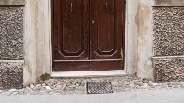 A double wooden door with a metal arch above it, set in a stone wall with a number '55' sign above. - In the ancient Jewish ghetto (Ferrara)