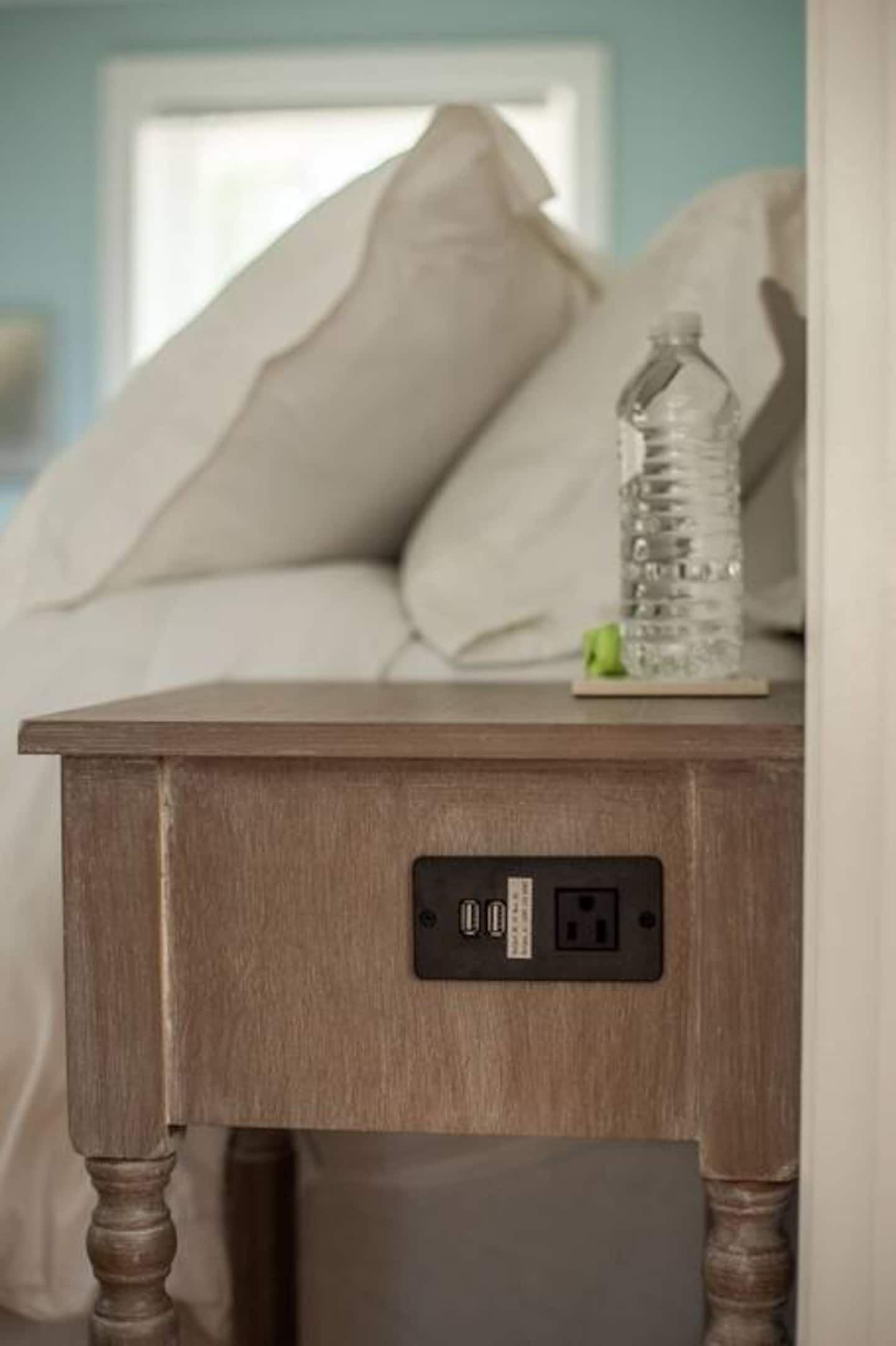 A wooden bedside table with a power outlet and USB ports, a water bottle, and a small plant on a bed with beige pillows.