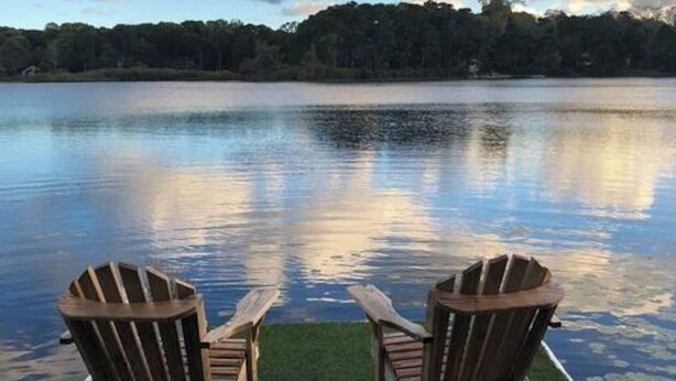 Dos sillas Adirondack mirando a un lago tranquilo con una orilla boscosa bajo un cielo parcialmente nublado.