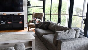 Una sala de estar moderna con un sofá gris, una mesa de centro y un televisor sobre un mueble negro. Hay un ventanal con vistas a zonas verdes.