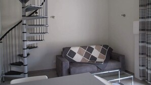 A modern room with a grey sofa, a small table, and a spiral staircase. - Gîte l'Amandier (Vogüé)