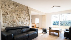 A living room with a stone wall, a black leather sofa, a wooden coffee table, and a view through large glass doors. - In nature, 3 bedroom house sleeps 8-10, totally renovated in 2019 (Aouste-sur-Sye)