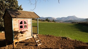 A wooden cabin with a red window and a small porch, set against a backdrop of a green field and mountains. - In nature, 3 bedroom house sleeps 8-10, totally renovated in 2019 (Aouste-sur-Sye)