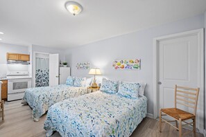 Ett sovrum med två sängar, en stol, ett nattduksbord, en lampa, en tavla och en liten köksdel.
