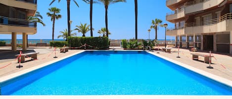 Ein rechteckiger Swimmingpool mit klarem, blauem Wasser, umgeben von Palmen und einem Gebäude mit Balkonen.