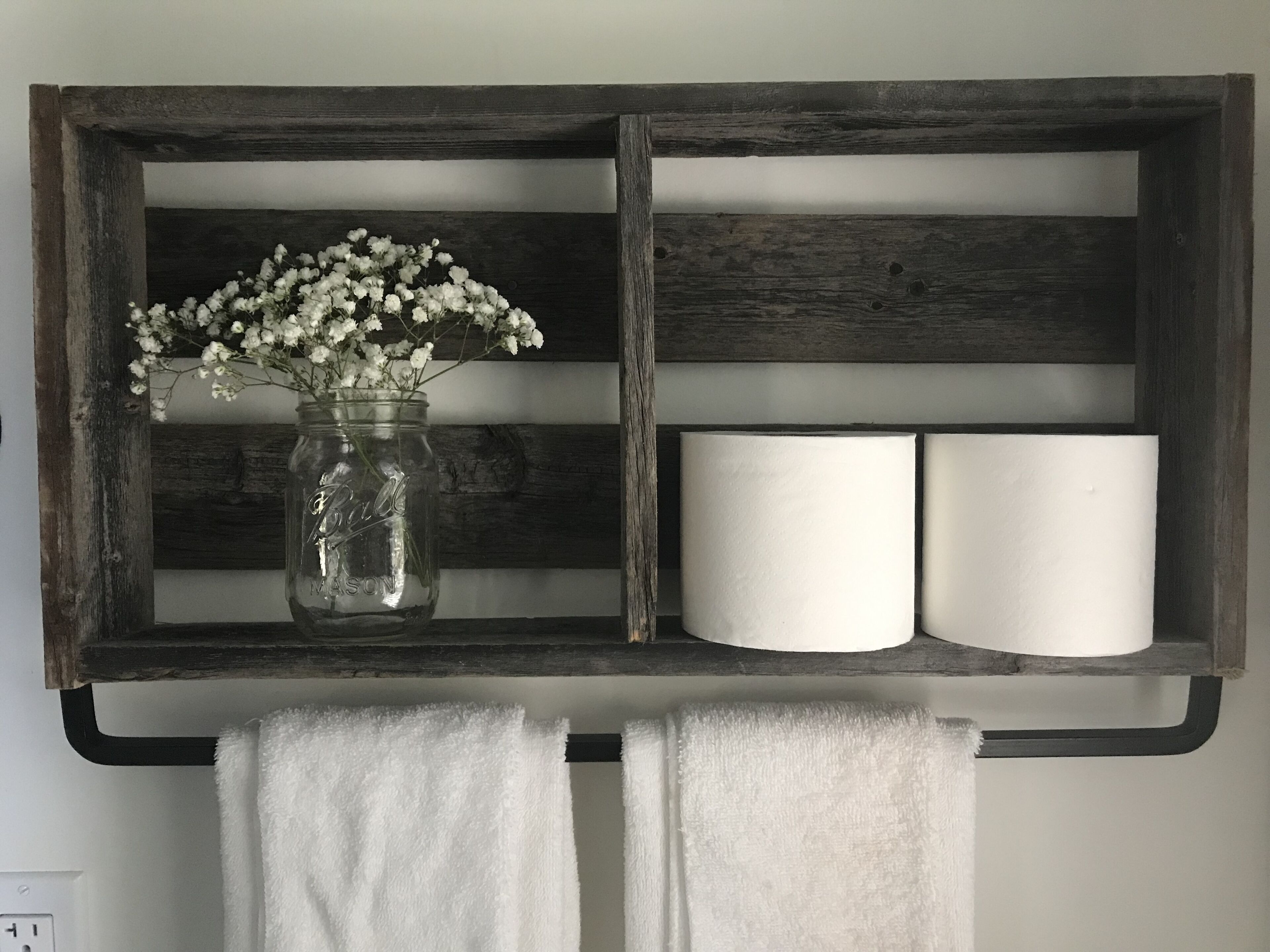 A rustic wooden shelf with a glass jar of white flowers, two white candles, and two white towels.