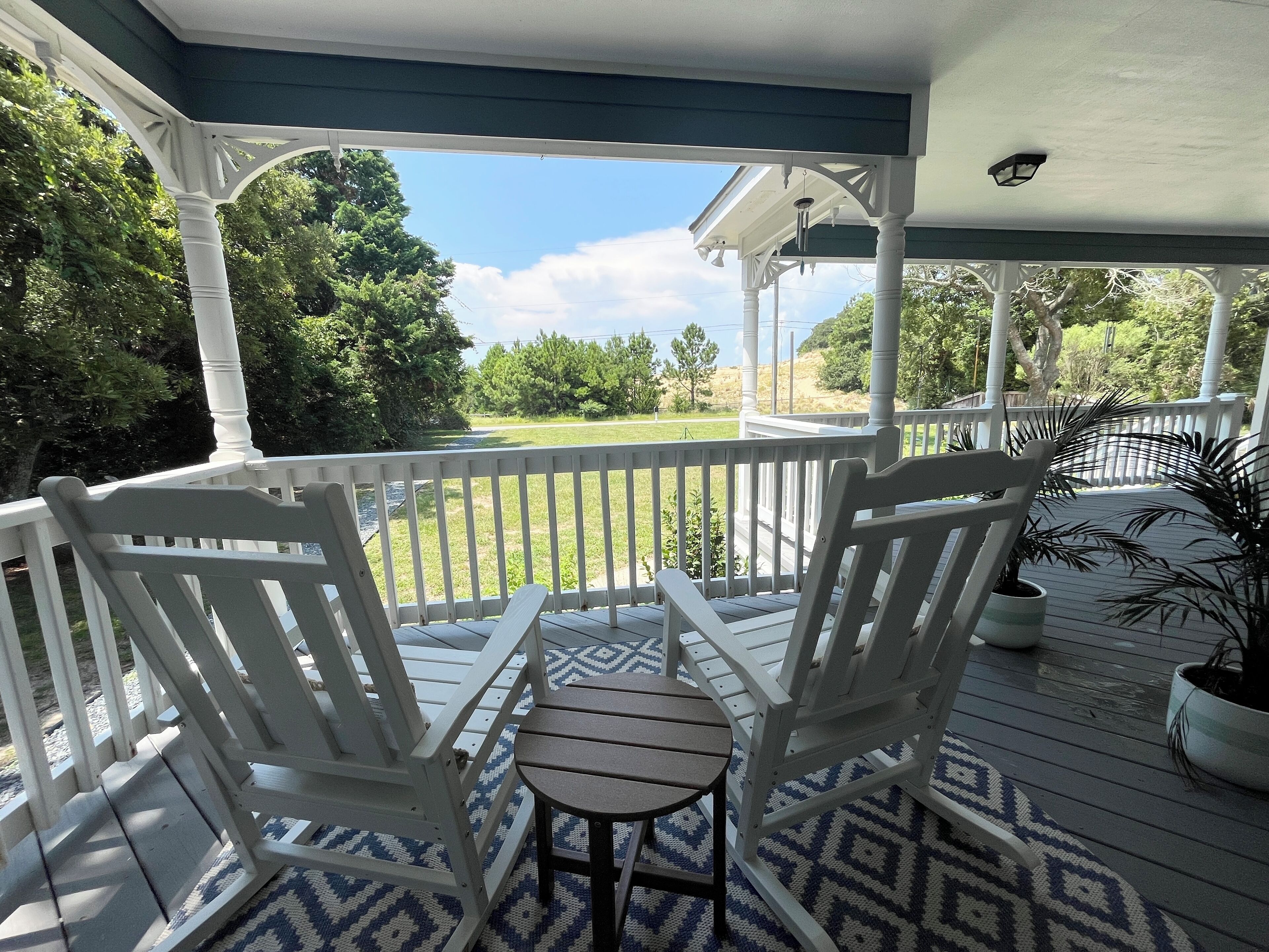 A covered porch with two white Adirondack chairs and a small table, overlooking a grassy area and trees.