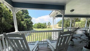 A covered porch with two white Adirondack chairs and a small table, overlooking a grassy area and trees. - On Jockey Ridge State Park + Hot Tub & Center of OBX (Nags Head)