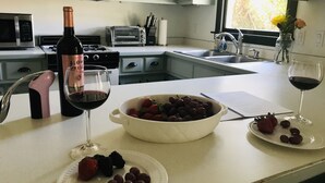 A kitchen with a white countertop, a wine bottle, two glasses of red wine, a bowl of grapes, and a plate with strawberries and blackberries. - On Jockey Ridge State Park + Hot Tub & Center of OBX (Nags Head)