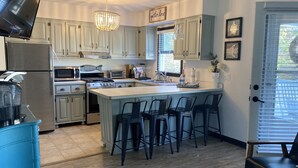 A modern kitchen with white cabinets, a central island with bar stools, stainless steel appliances, and a chandelier. - On Jockey Ridge State Park + Hot Tub & Center of OBX (Nags Head)