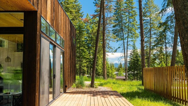 Un edificio moderno de madera con amplios ventanales, una terraza de madera y un paisaje boscoso de fondo.