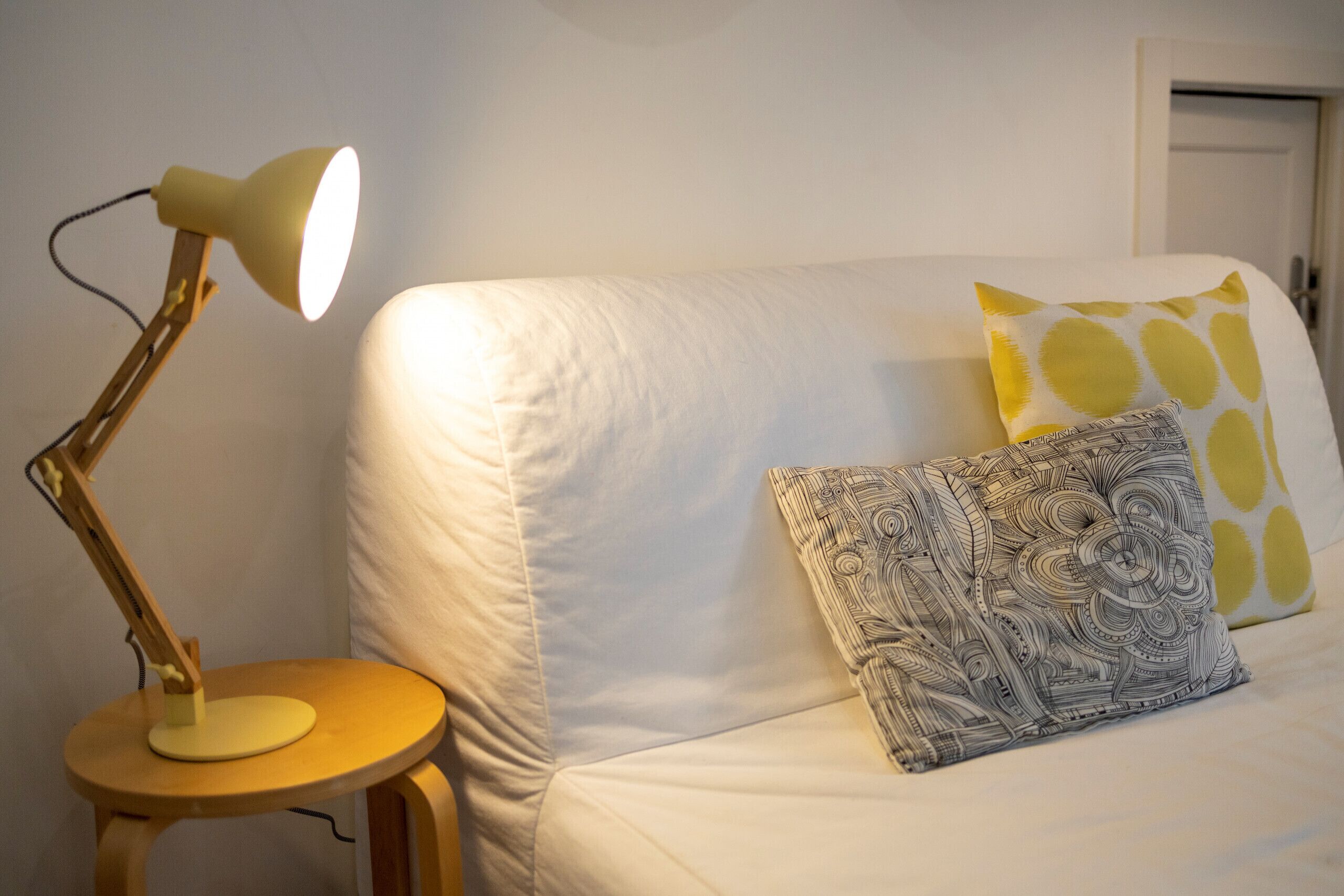A bed with a white mattress, a patterned pillow, and a yellow pillow with a circular pattern. There is a wooden nightstand with a lamp to the left of the bed.