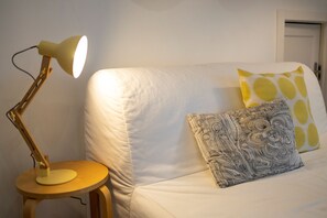 Ein Bett mit weißer Matratze, einem gemusterten Kopfkissen und einem gelben Kopfkissen mit kreisförmigem Muster. Links neben dem Bett steht ein Nachttisch mit einer Lampe.