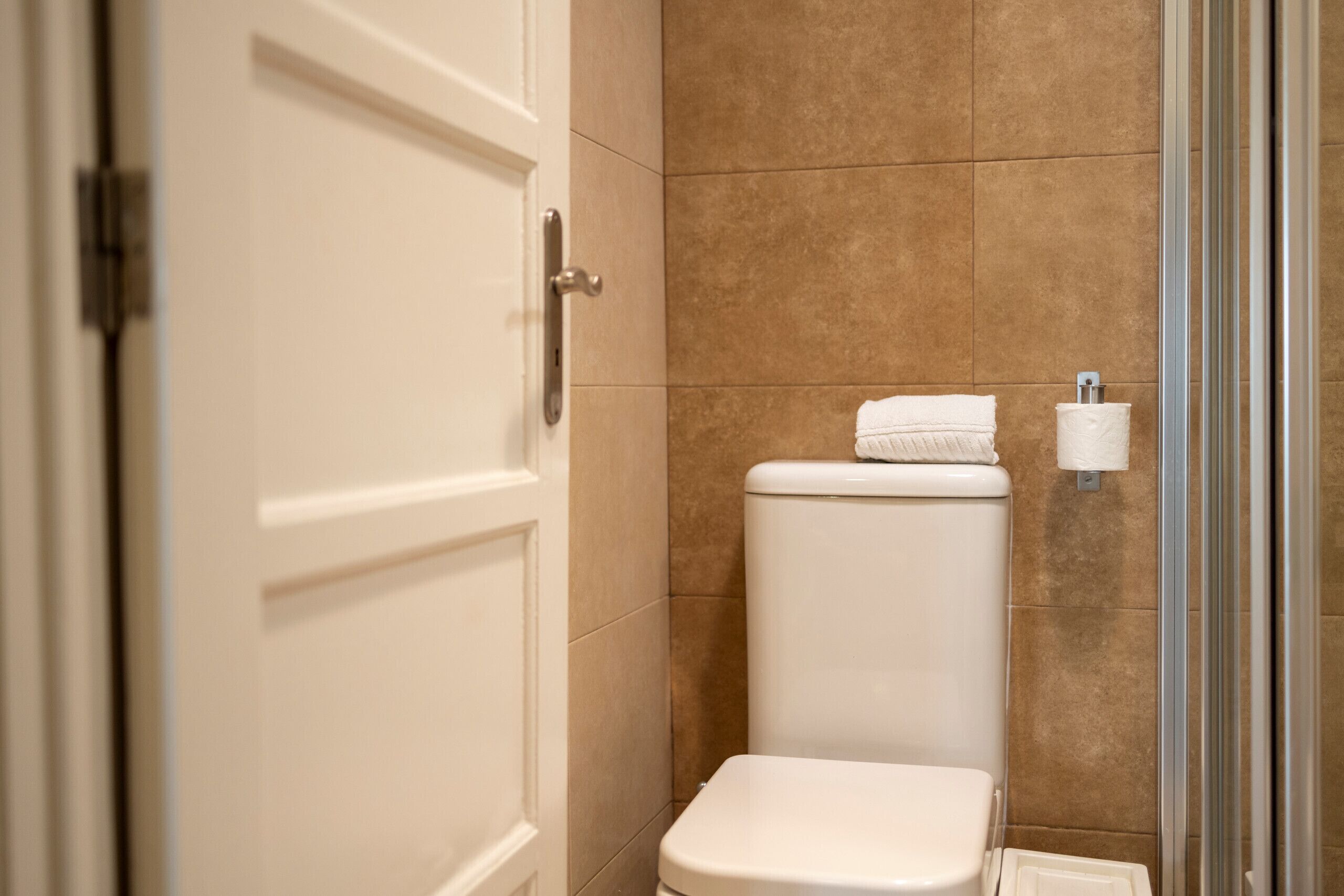 Un baño con inodoro blanco, baldosas color beige y una toalla blanca sobre el tanque.