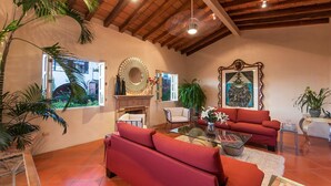 A spacious living room with red sofas, a glass coffee table, and a fireplace. There are multiple windows allowing natural light, and the room has a wooden ceiling.