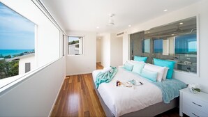 A bedroom with a large window, a bed with multiple pillows, a bedside table, and a view of the outdoors. - Oleander Holiday Home (Airlie Beach)
