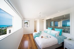 Ein Schlafzimmer mit großem Fenster, einem Bett mit vielen Kissen, einem Nachttisch und Blick ins Freie.