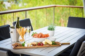 A table set with seafood, bread, cheese, and a bottle of champagne on an outdoor patio with a view of greenery. - Oleander Holiday Home (Airlie Beach)