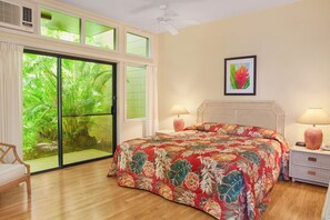 A room with a bed, two bedside lamps, a ceiling fan, a window with a view of greenery, and a framed picture on the wall.