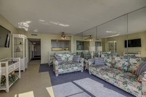Un salon avec deux canapés à motifs floraux, une table basse et une table à manger avec des chaises. On peut apercevoir une cuisine à travers la porte vitrée, un téléviseur fixé au mur et un ventilateur de plafond.