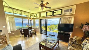 A spacious living room with a large TV, comfortable seating, a dining area, and a balcony with a view. - Kapalua Ridge 411- Dazzling Ocean & Island Views! (Lahaina)