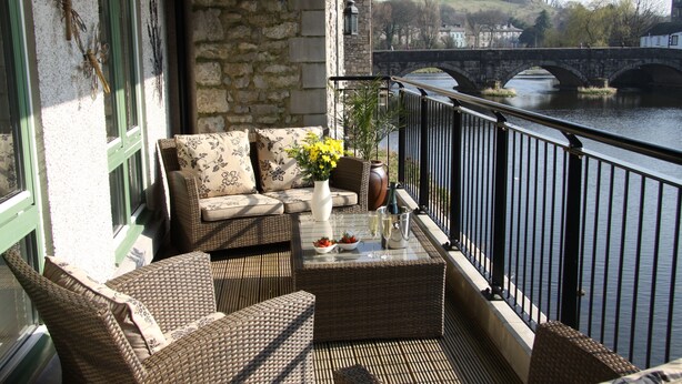 Un balcone con mobili in vimini, un vaso con fiori e vista su un fiume e un ponte.